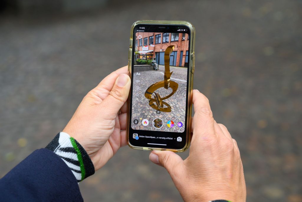 A person holding a smartphone displaying an augmented reality sculpture by Vira DG on the screen. The background shows a cobblestone street and buildings.