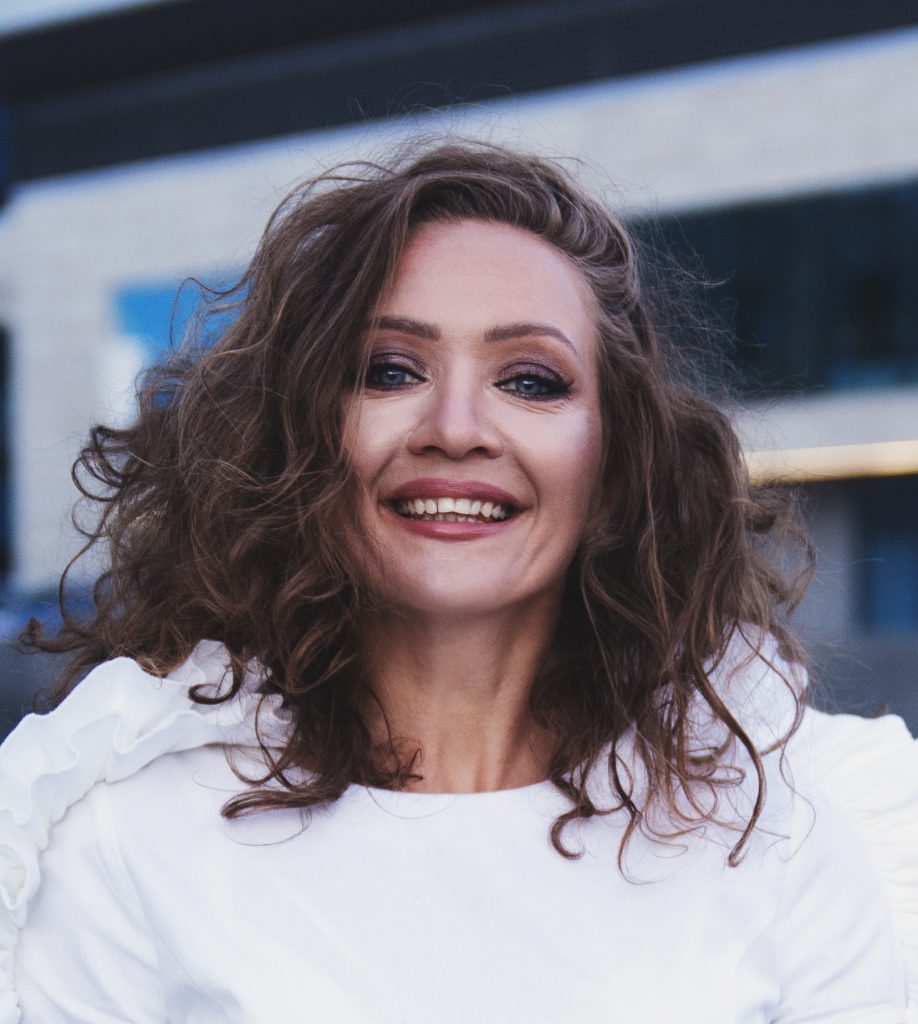 Smiling portrait of Vira DG, a Ukrainian artist, with curly hair, wearing a white top, standing outdoors against a modern architectural background.